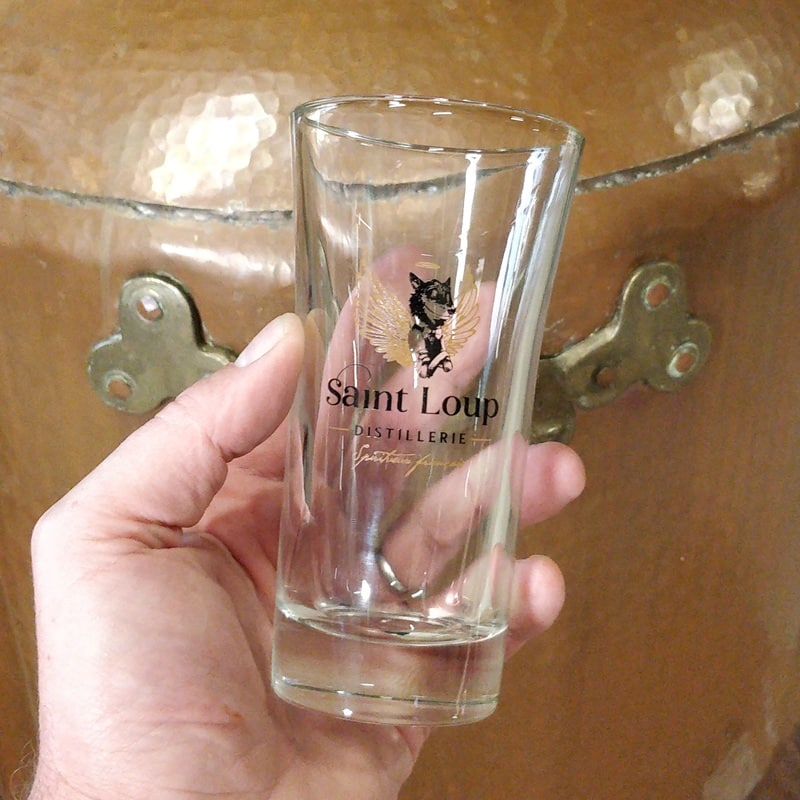 Verre long drink avec le logo de la distillerie saint loup devant un alambic en cuivre